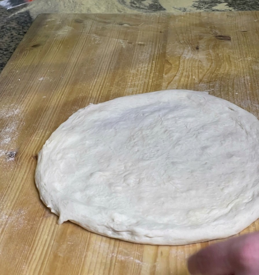 Uno chef che stende uno strato di salsa di pomodoro rossa vibrante al centro di un impasto per pizza crudo su un tagliere di legno.
