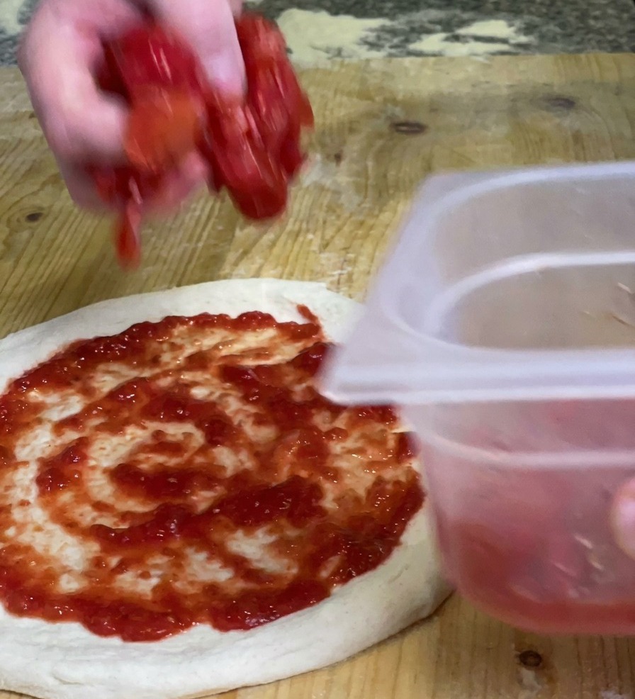 Una mano che distribuisce pezzi di polpa di pomodoro densa su una base pizza già ricoperta di salsa rossa.