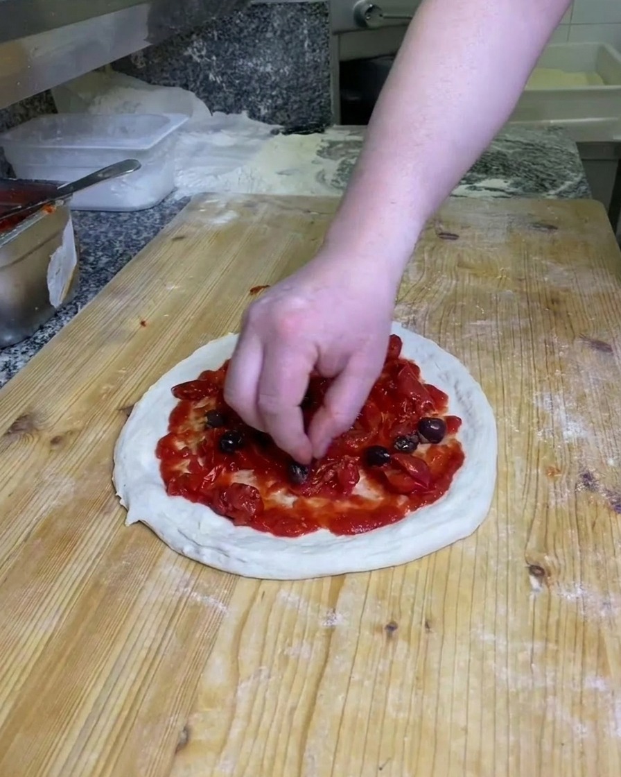 Una mano che posiziona olive nere intere sopra limpasto della pizza coperto di pomodoro su una superficie di legno infarinata.