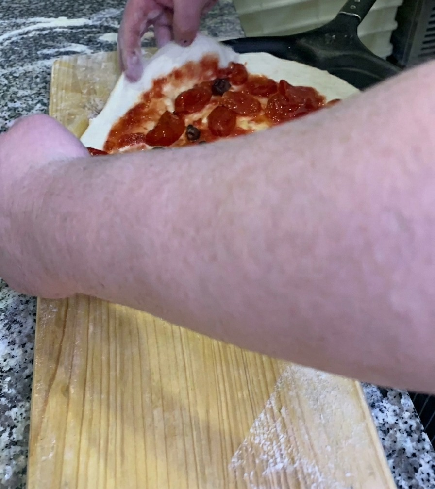 Lo chef che fa scivolare una pizza cruda e condita su una pala di metallo nera dal banco di lavoro in legno.