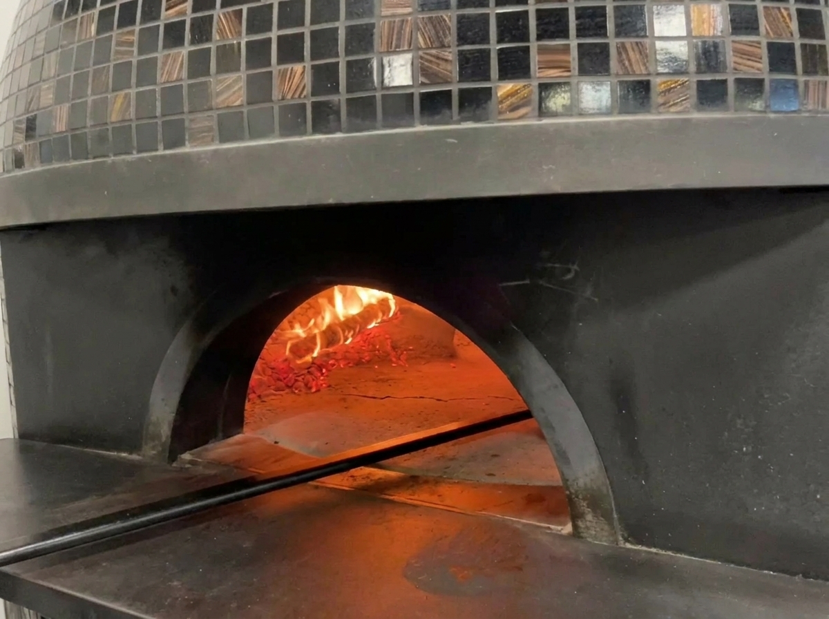 Vista allinterno di un tradizionale forno a legna piastrellato con fiamme che lambiscono la cupola e una pizza in cottura.