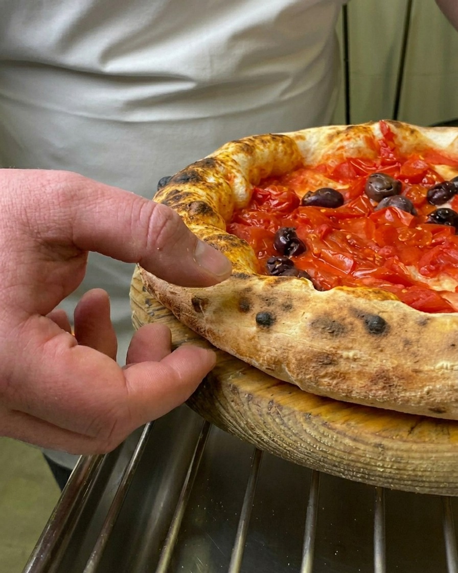 Il dito di uno chef che preme sul cornicione soffice, brunito e alveolato di una pizza Marinara appena sfornata.