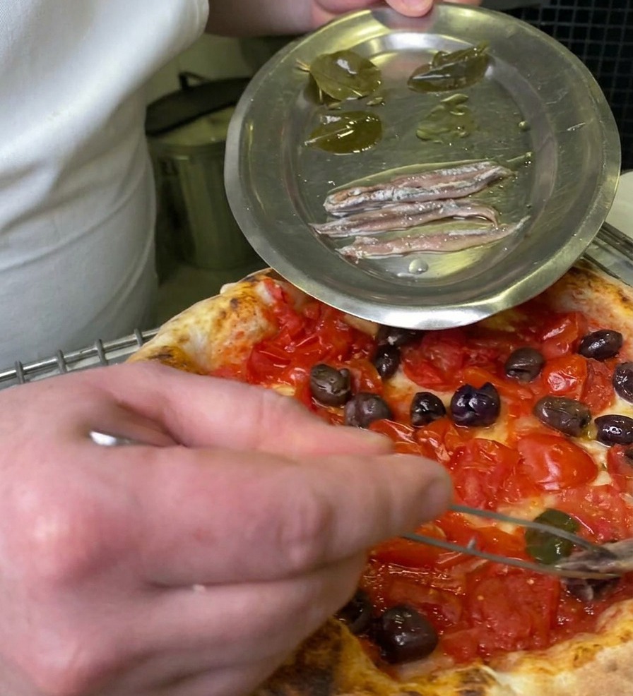 Uno chef che usa delle pinzette per prelevare una foglia di alloro in salamoia da un piatto di metallo e posizionarla sulla pizza.