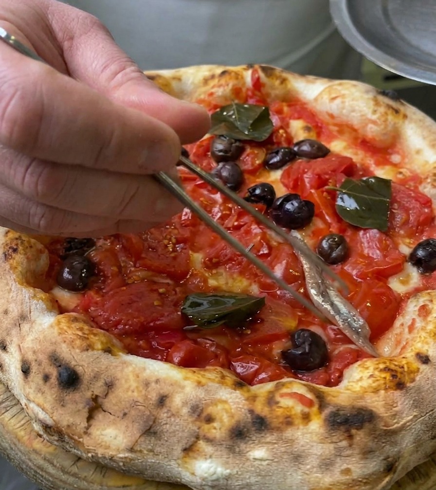 Primo piano di un filetto di acciuga argentata che viene adagiato sul cornicione della pizza con pinzette culinarie.