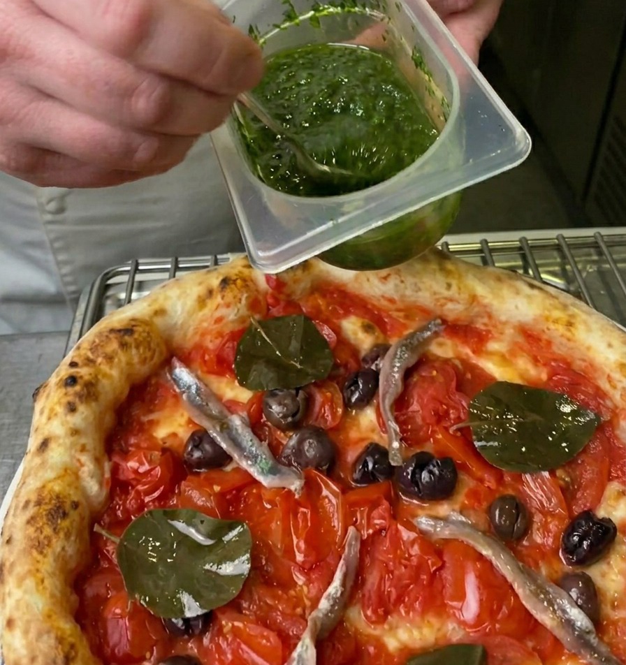 Uno chef che versa una salsa verde brillante da un contenitore sulla pizza usando un cucchiaino.