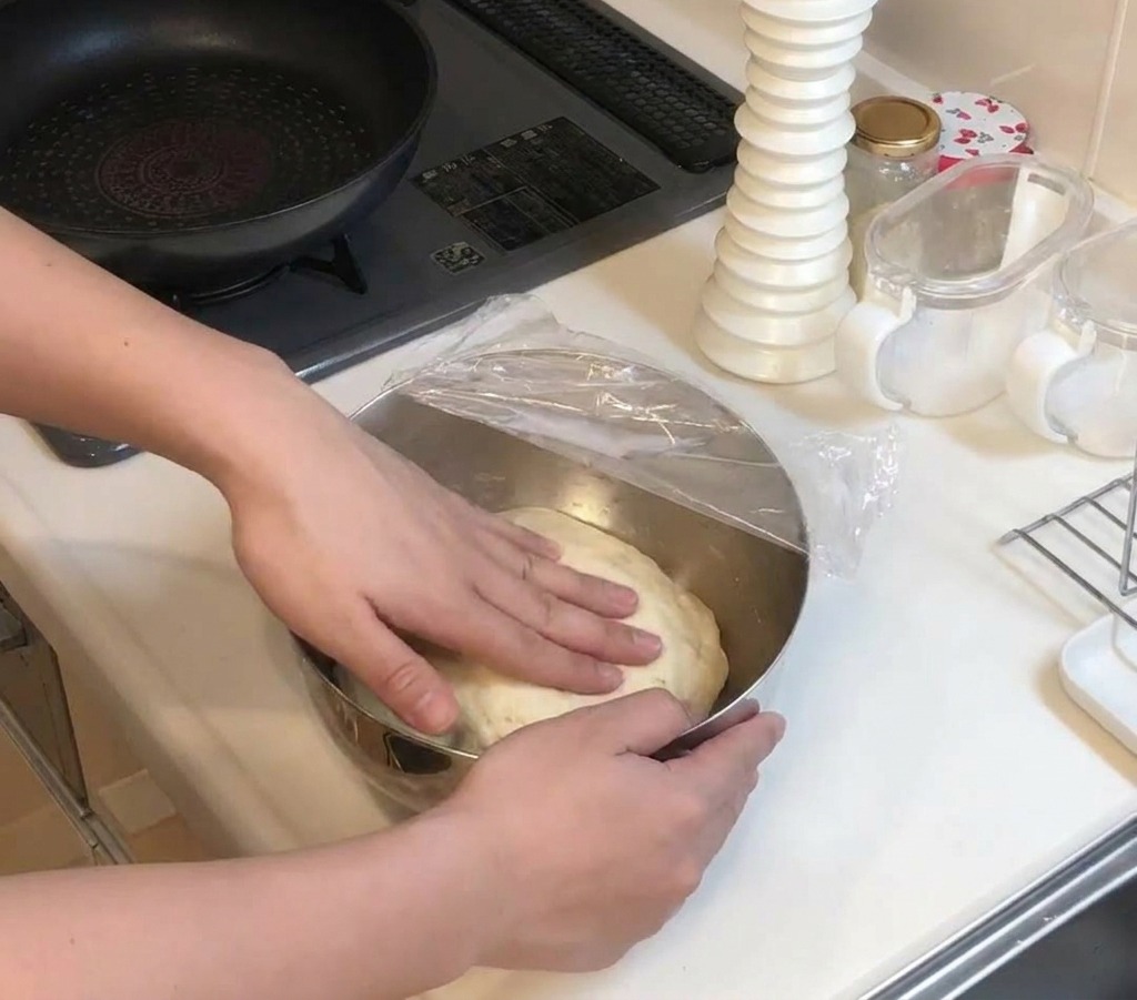 Mani che premono su un panetto di impasto lievitato e chiaro dentro una ciotola coperta da pellicola trasparente.