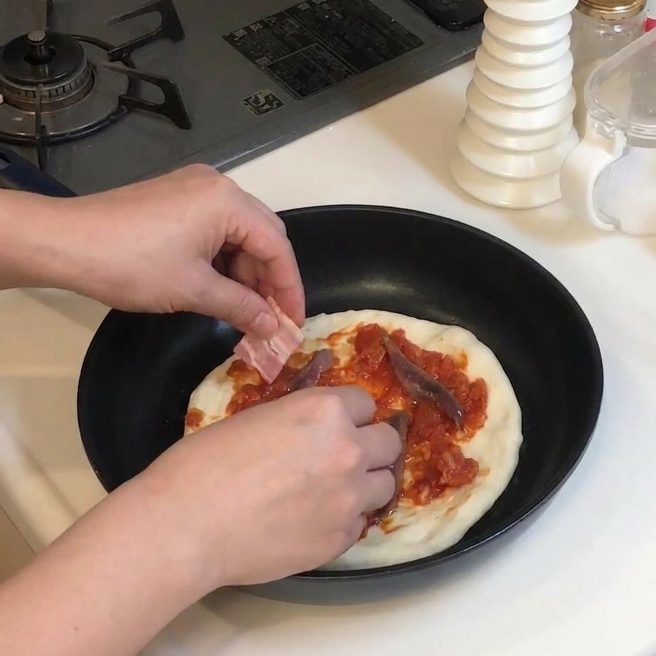 Mani che posizionano fette rettangolari di pancetta cruda su una pizza già condita con pomodoro e acciughe.