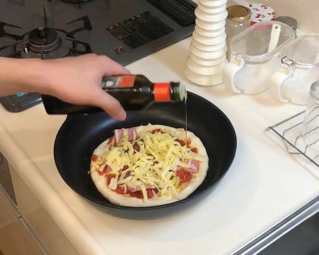 Primo piano di una mano che versa olio da una bottiglia scura su una pizza preparata in padella con formaggio e pancetta.
