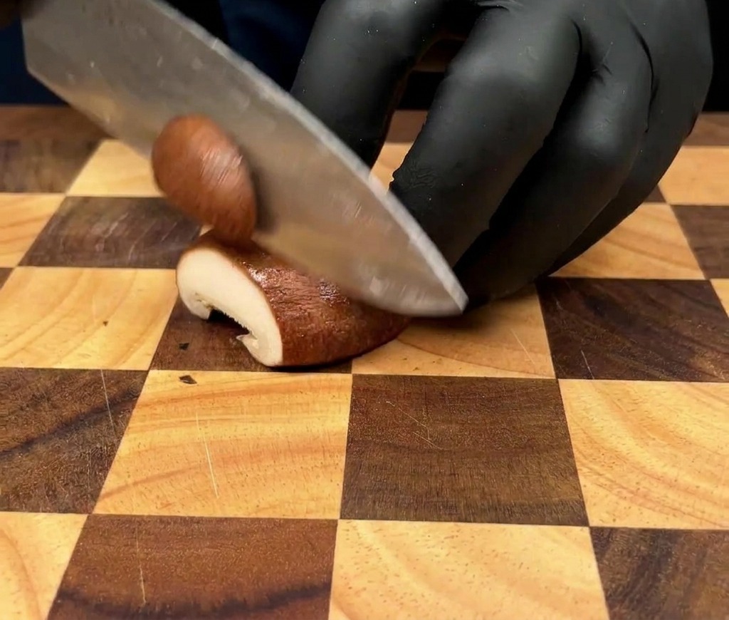 Un primo piano di una persona che indossa guanti neri e affetta un fungo cardoncello a fettine sottili su un tagliere di legno a scacchi.