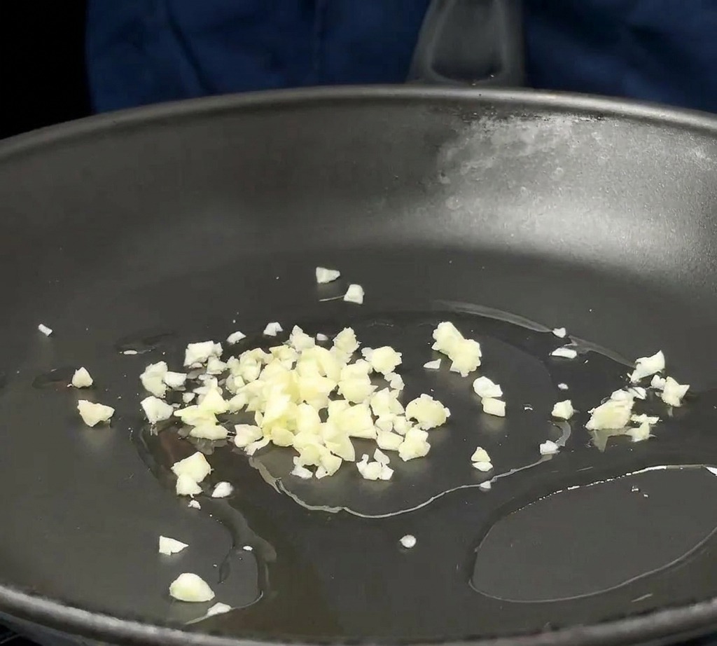 Aglio tritato e cipolle rosse a cubetti sfrigolano in un sottile strato dolio sul fondo di una padella scura.