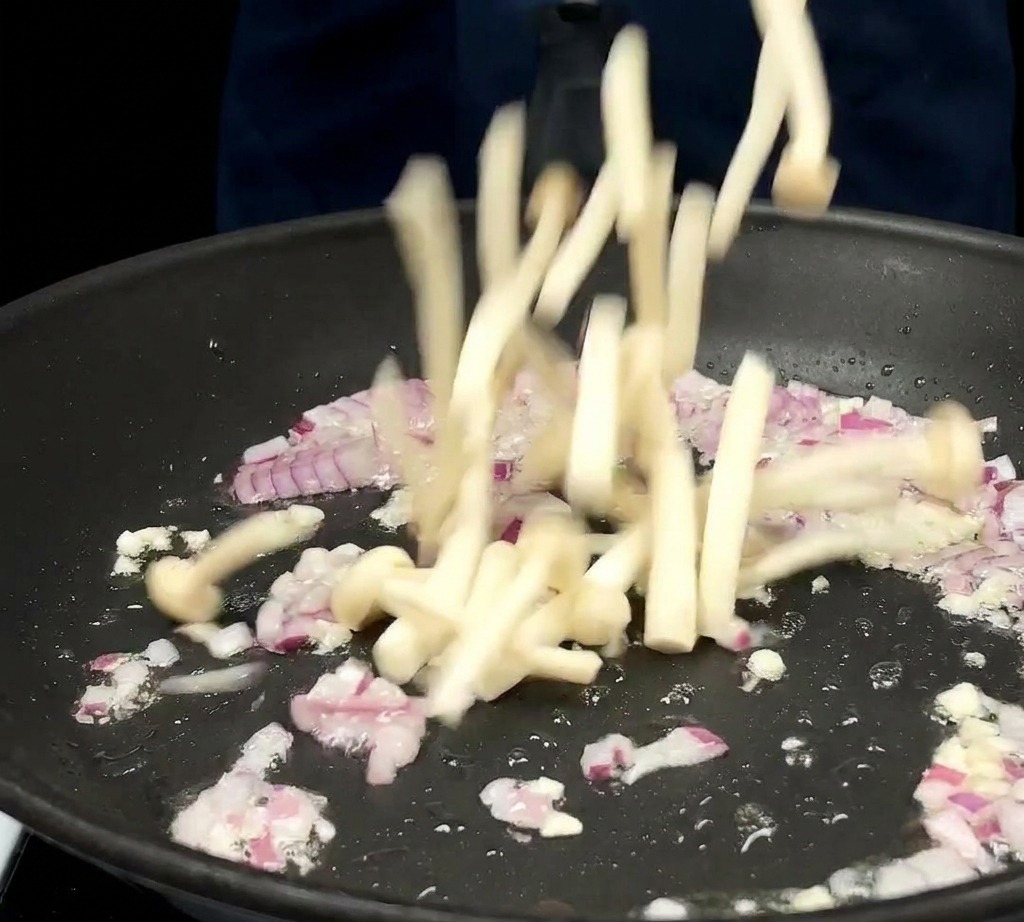 Un mazzetto di lunghi funghi shimeji bianchi viene fatto cadere in una padella contenente cipolle rosse e aglio soffritti.