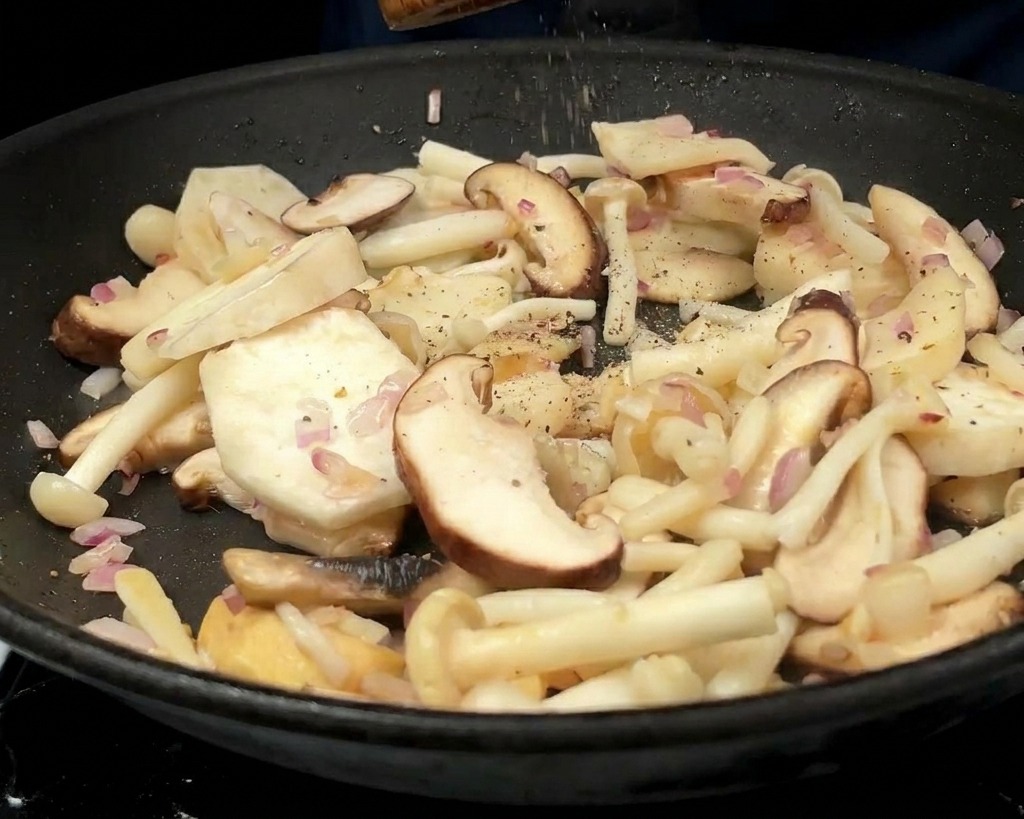 Una persona usa un macinapepe di legno per macinare pepe nero su una varietà di funghi e cipolle saltati in padella.
