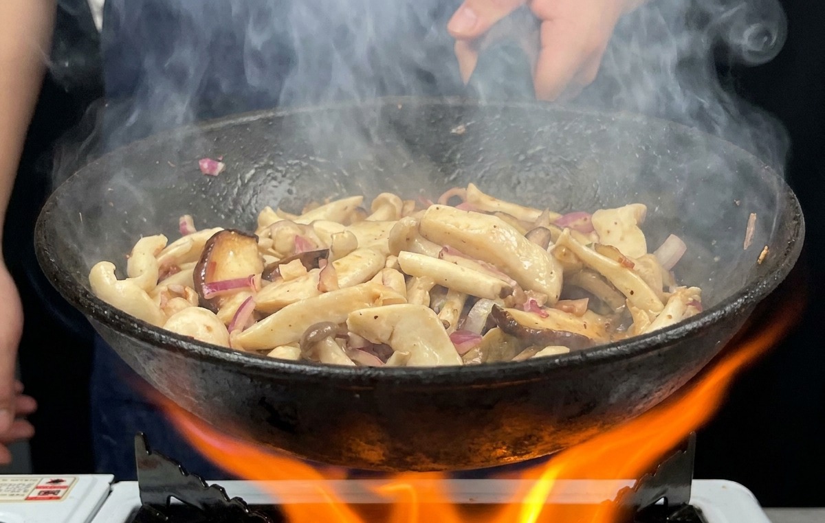 Una padella piena di funghi viene saltata su una grande e vivace fiamma arancione con il vapore che sale verso lalto.