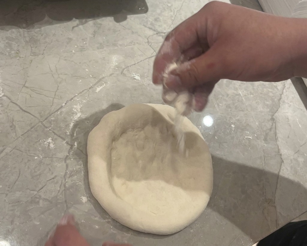 Mani che premono e stendono un panetto di impasto crudo su un piano di marmo grigio infarinato.