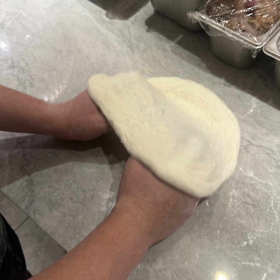 Mani che sollevano e ruotano limpasto della pizza per stenderlo in forma circolare con un bordo esterno visibile.