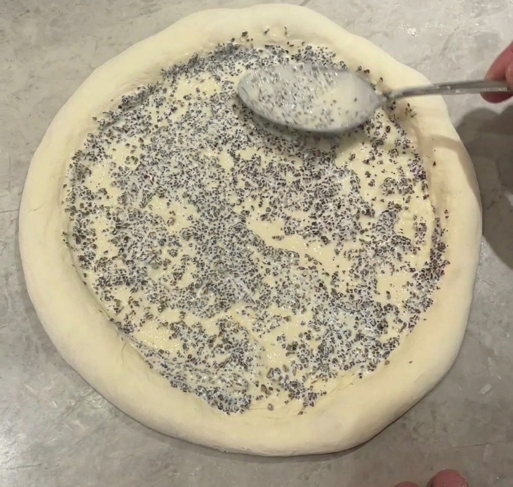 Un cucchiaio di metallo che spalma una densa salsa al tartufo nero sul centro di una base per pizza cruda.