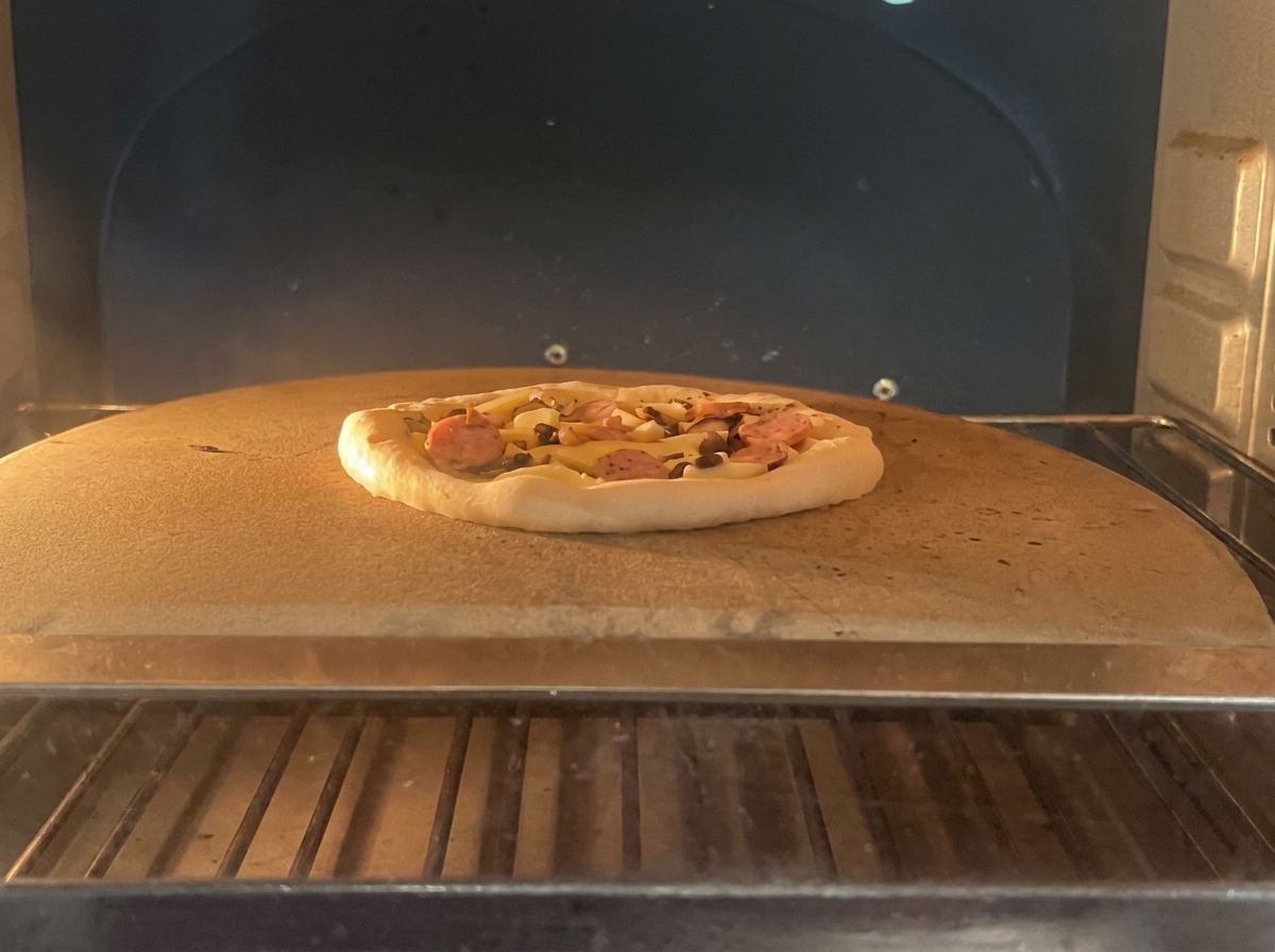 Una pizza non cotta con salame e funghi appoggiata su una pietra refrattaria allinterno di un forno professionale.