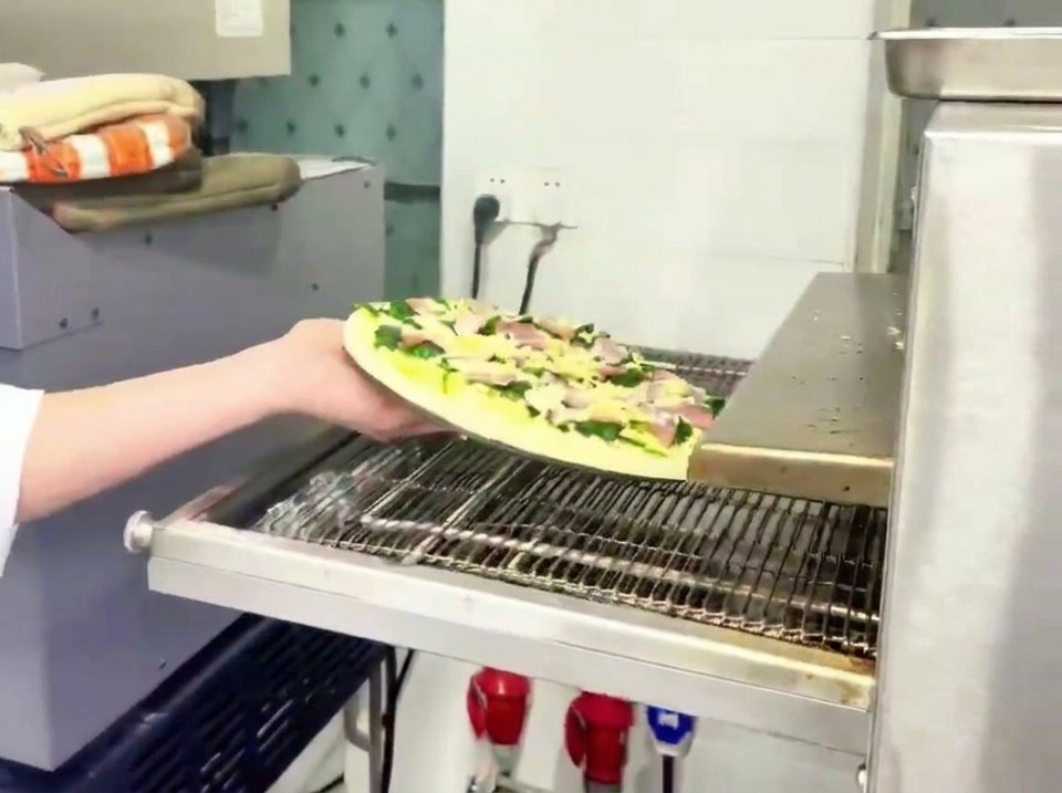 Addetto che inserisce la pizza con spinaci e pancetta su un nastro trasportatore di un forno professionale.