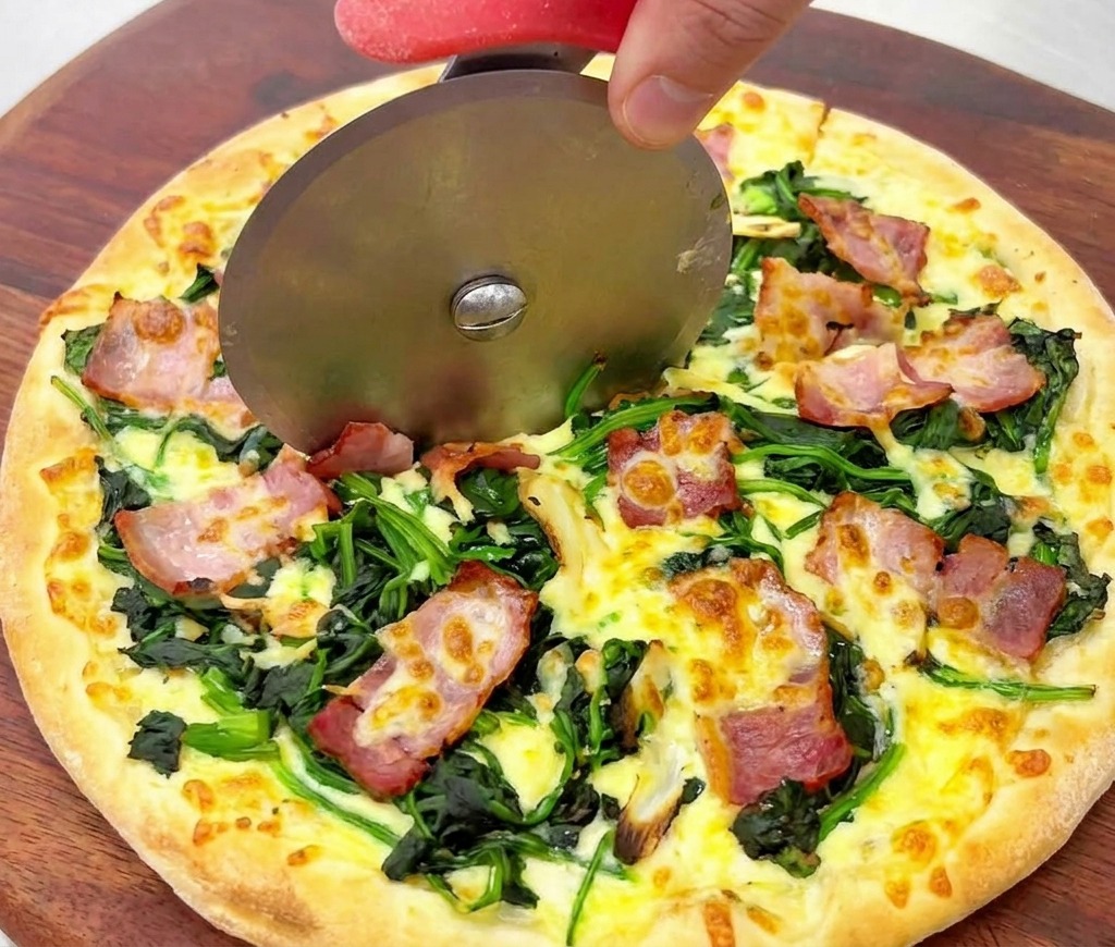 Primo piano di una rotella per pizza che taglia una pizza dorata con spinaci e pancetta croccante.