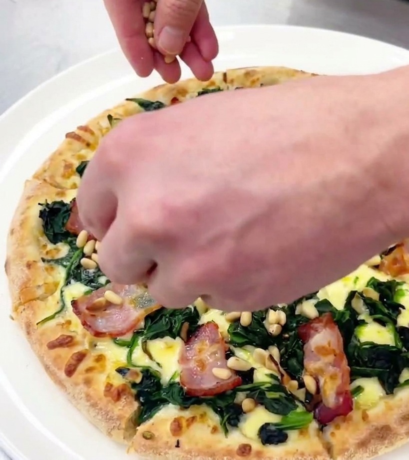 Mano che sparge pinoli tostati su una pizza appena tagliata su un piatto bianco.