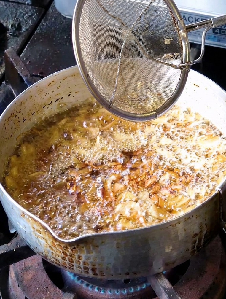 Un colino a maglia fine usato per raccogliere fettine daglio fritte di colore marrone dorato da un pentolino di olio bollente.
