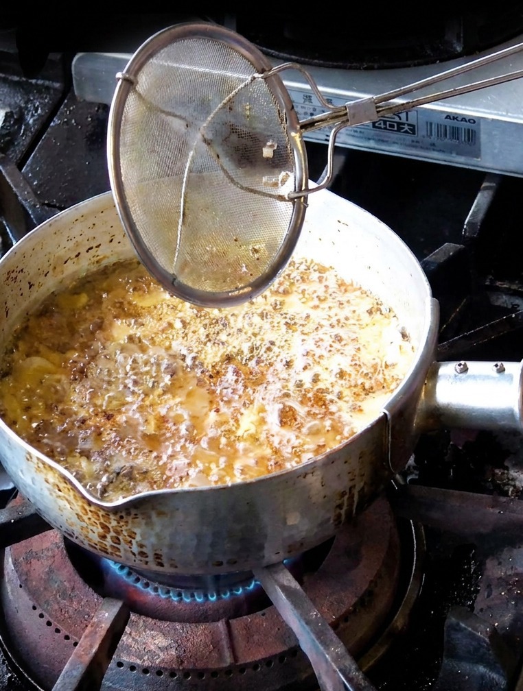 Una mano che usa un colino per rimuovere fettine daglio fritte di colore marrone medio da un pentolino di olio scuro.