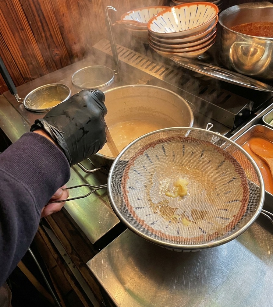 Brodo bianco e cremoso versato attraverso un colino metallico in una ciotola di ramen decorata.