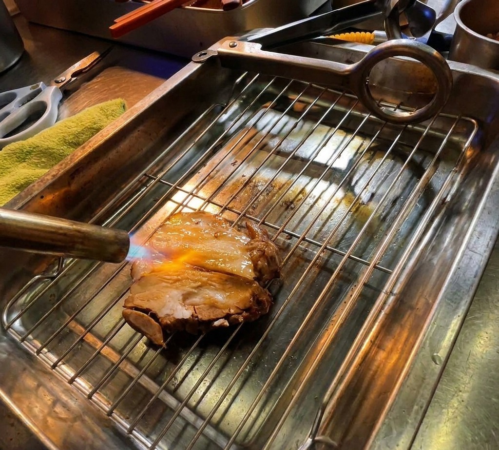 Un cannello da cucina che scotta una fetta spessa di maiale chashu su una griglia metallica con fiamme visibili.