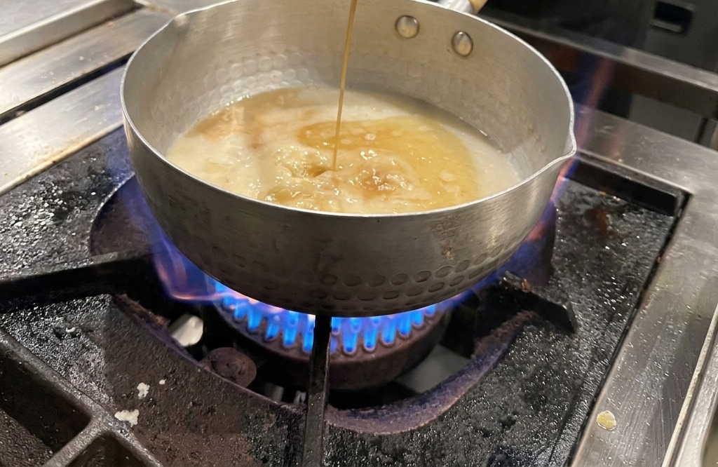 Salsa di soia scura versata da un mestolo in una pentola con brodo chiaro su un fornello acceso.