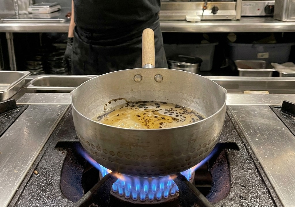 Gocce e vortici di olio nero che galleggiano sopra una salsa densa marrone chiaro in una pentola di metallo martellato.