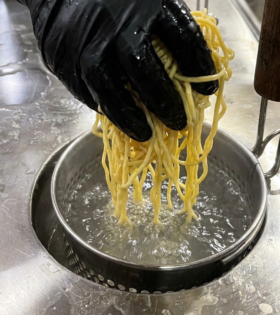 Una mano abbassa un colino metallico pieno di noodles gialli nellacqua bollente.