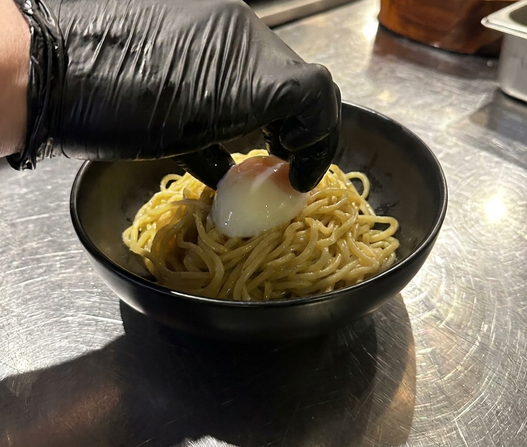 Una mano posiziona delicatamente un uovo onsen intero sopra una porzione di ramen in una ciotola nera.