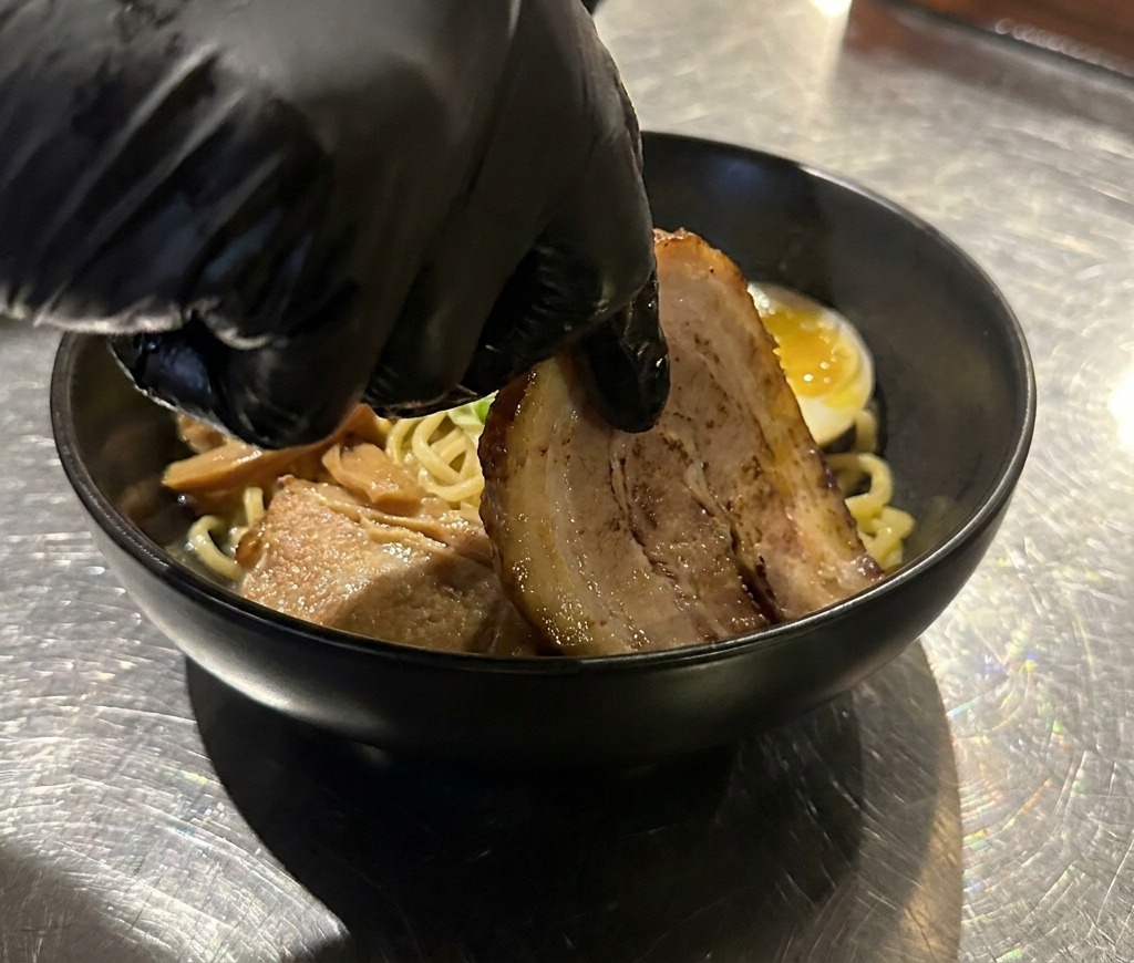 Una mano che posiziona una fetta di chashu di maiale tenero e scottato in una ciotola di noodles già guarnita.