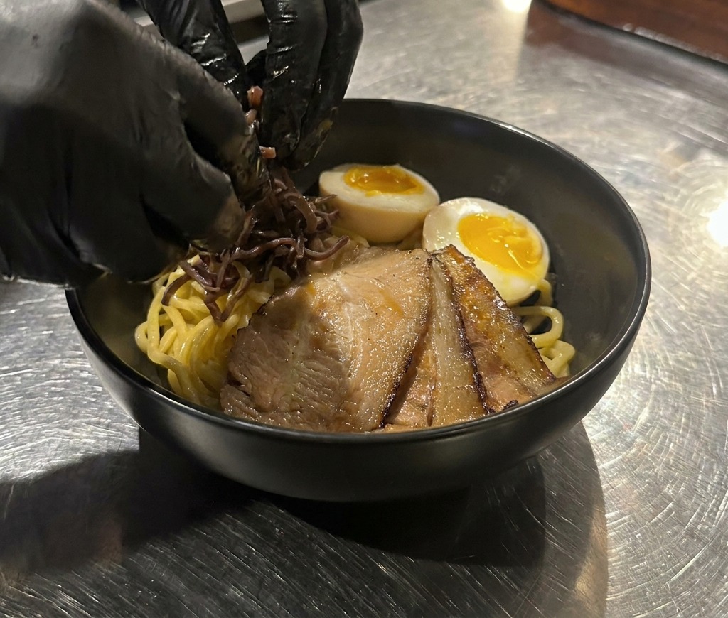 Mani che posizionano strisce di funghi wood ear scuri su una ciotola di noodles con chashu e uovo.