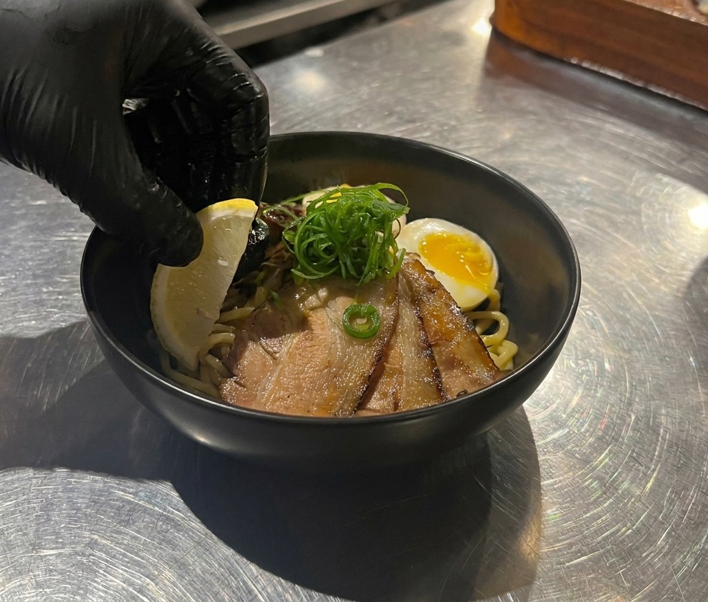 Una mano posiziona uno spicchio di limone giallo brillante in una ciotola nera con noodles, chashu, uovo e cipollotti.