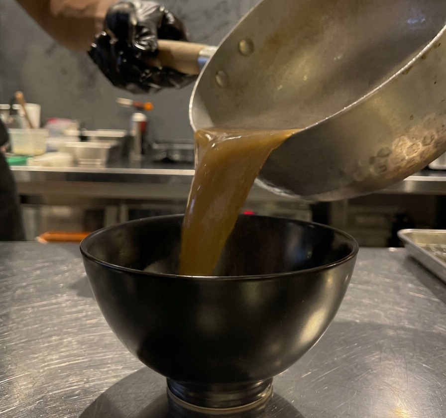 Salsa densa marrone allaglio nero versata da un pentolino in una piccola ciotola di ceramica nera.