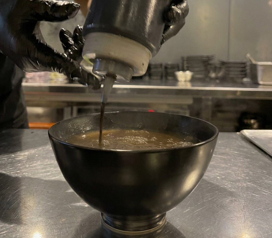 Olio nero spremuto da un flacone in una ciotola di salsa marrone, creando gocce scure in superficie.