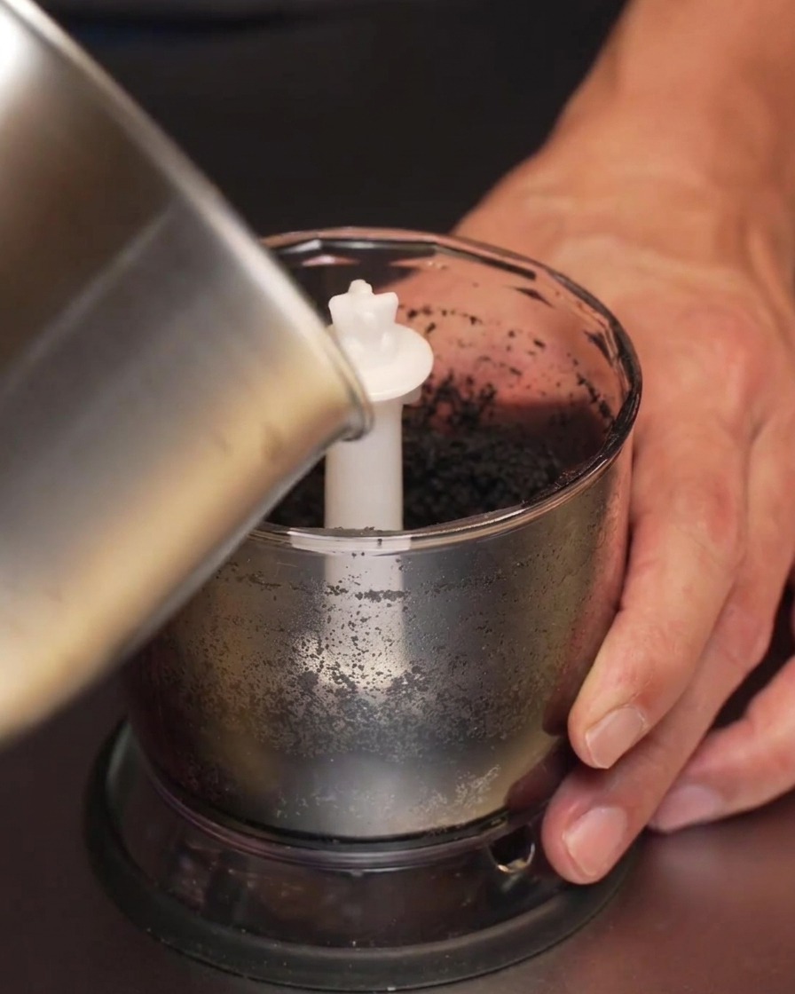 Olio infuso allaglio scurito versato da un pentolino di metallo in un boccale per frullatore contenente pasta daglio nero.