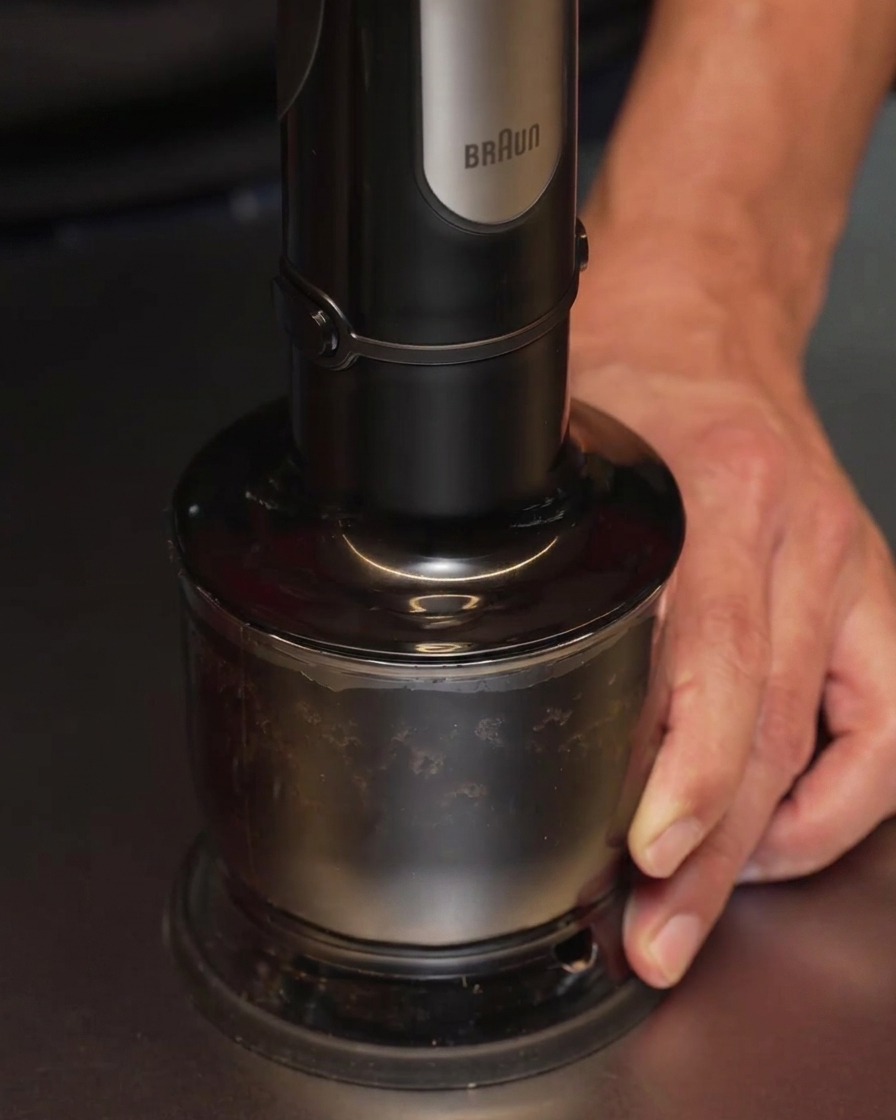 Un frullatore a immersione utilizzato per lavorare aglio carbonizzato e olio in un contenitore, trasformando il composto in un liquido nero liscio e opaco.