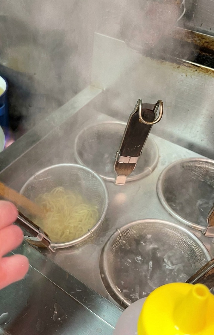 Noodle in cottura dentro un colino a rete immerso in una pentola di acqua che bolle in una cucina professionale.