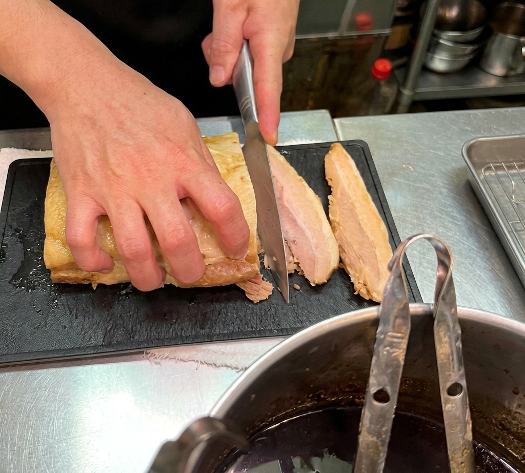 Primo piano di uno chef che taglia un pezzo di pancetta di maiale brasata in fette spesse su un tagliere nero.