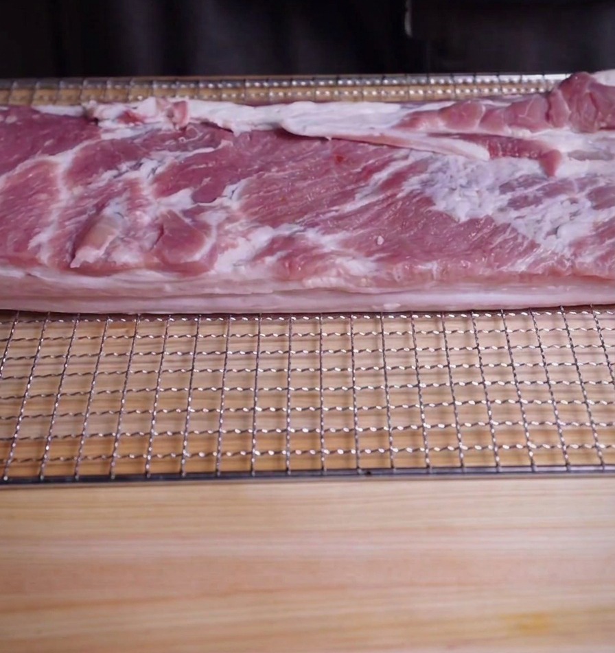 Un pezzo rettangolare di pancetta di maiale cruda su una griglia sopra un tagliere di legno.