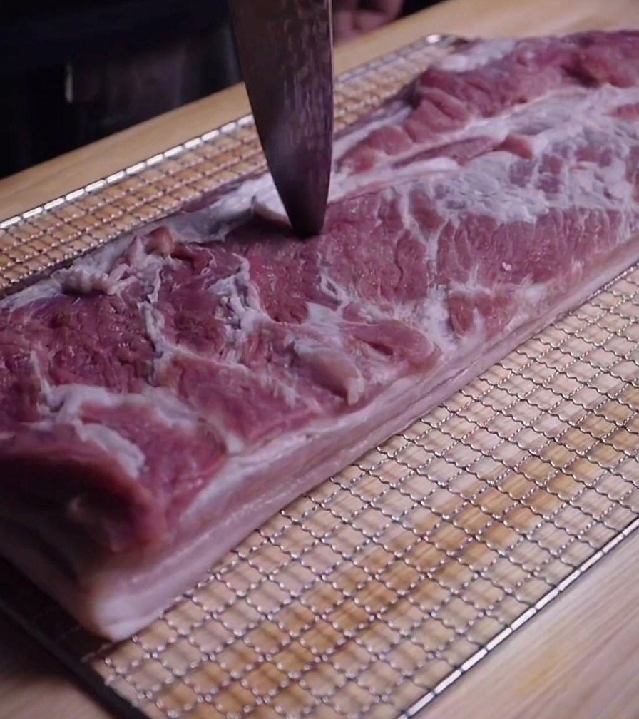 Dettaglio della punta di un coltello che pratica piccoli fori sulla superficie della pancetta cruda.
