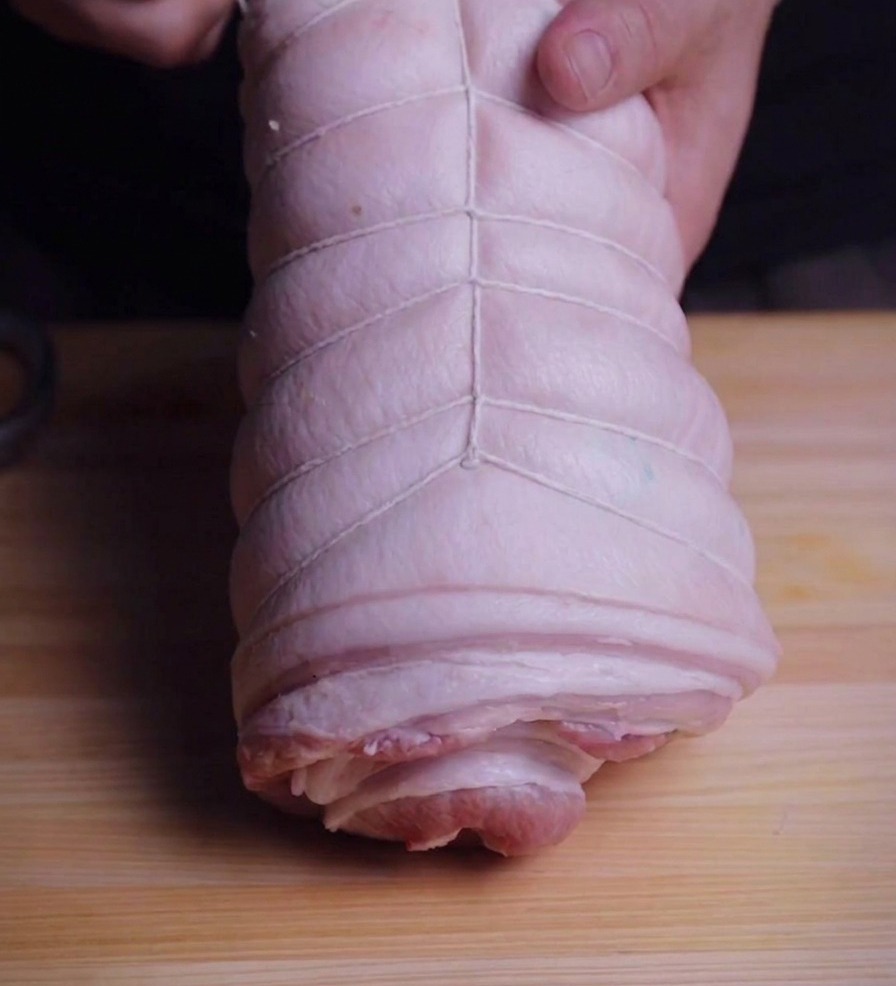 Mani che avvolgono un rotolo di pancetta di maiale con spago da cucina bianco su un tagliere.