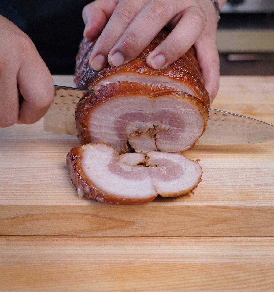 Uno chef che taglia fette tonde e spesse da un rotolo di maiale chashu freddo con un coltello affilato.