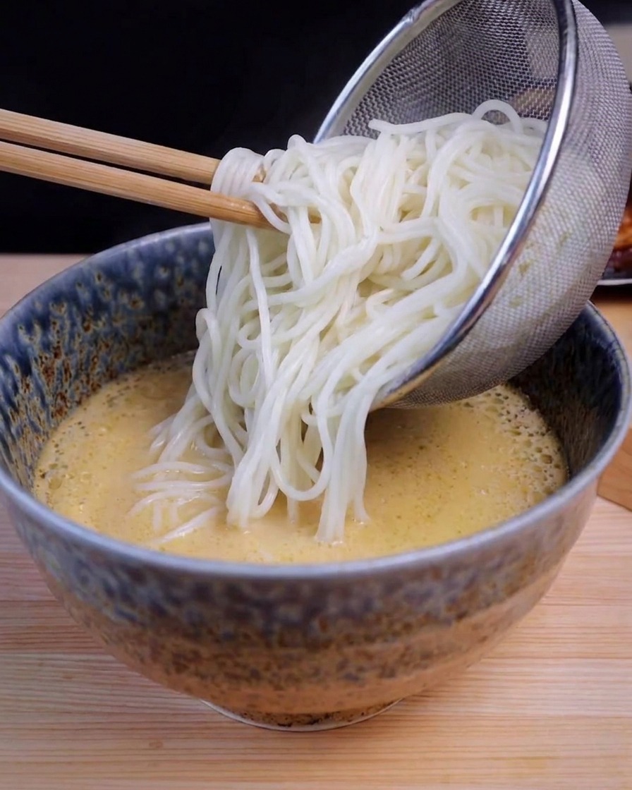 Un colino in acciaio che versa noodle bianchi in una ciotola azzurra con brodo giallo.