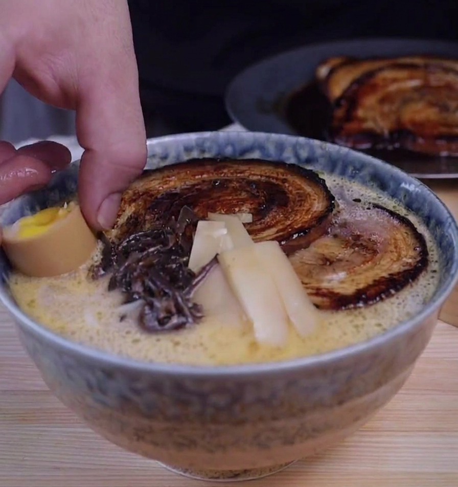 Una mano che aggiunge un uovo sodo tagliato a metà in una ciotola di ramen guarnita con chashu e funghi.