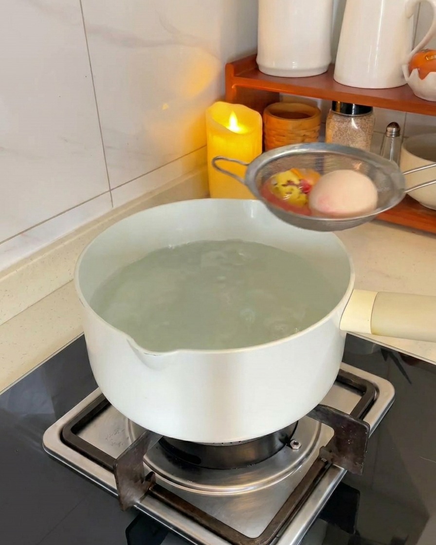 Due uova bianche in un colino a rete metallica che vengono immerse in una pentola di acqua bollente sul fornello.