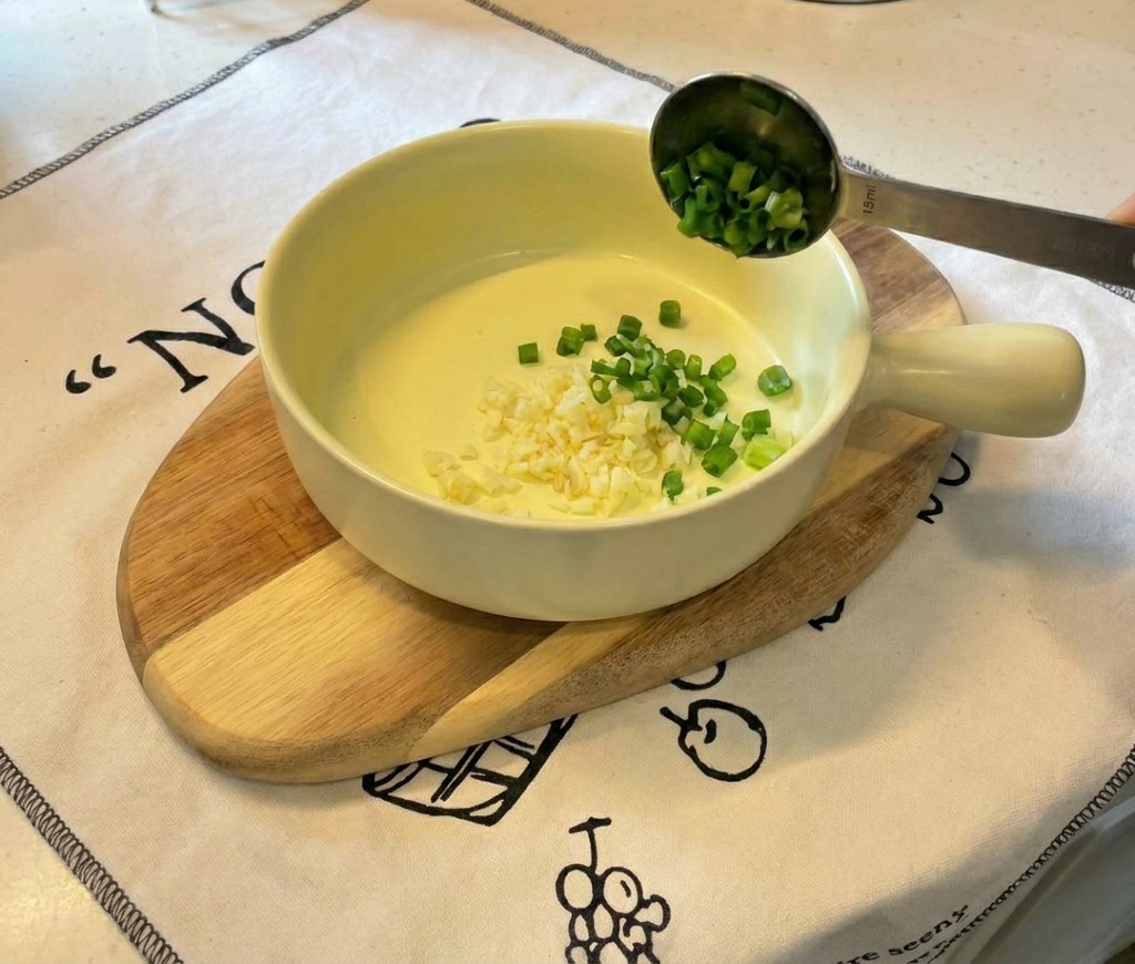 Una piccola ciotola in ceramica gialla su un tagliere di legno con aglio tritato e cipollotti affettati.