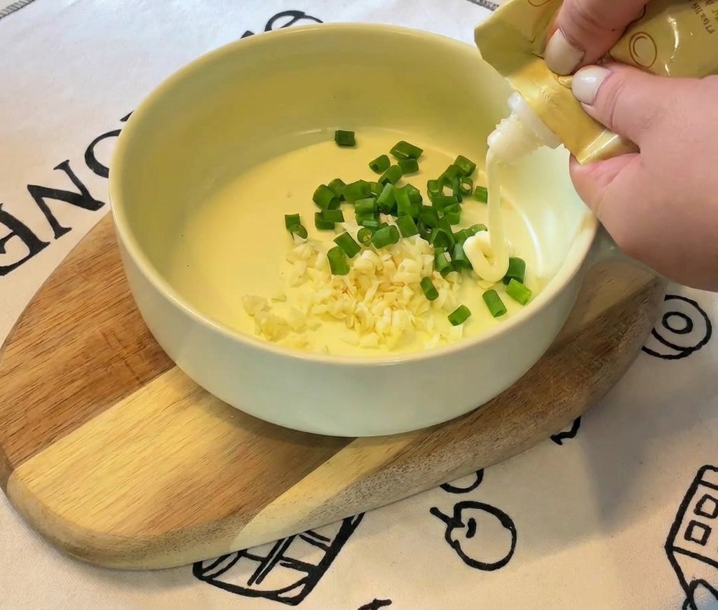 Mani che spremono maionese bianca da un tubetto giallo in una ciotola con aglio e cipollotto.